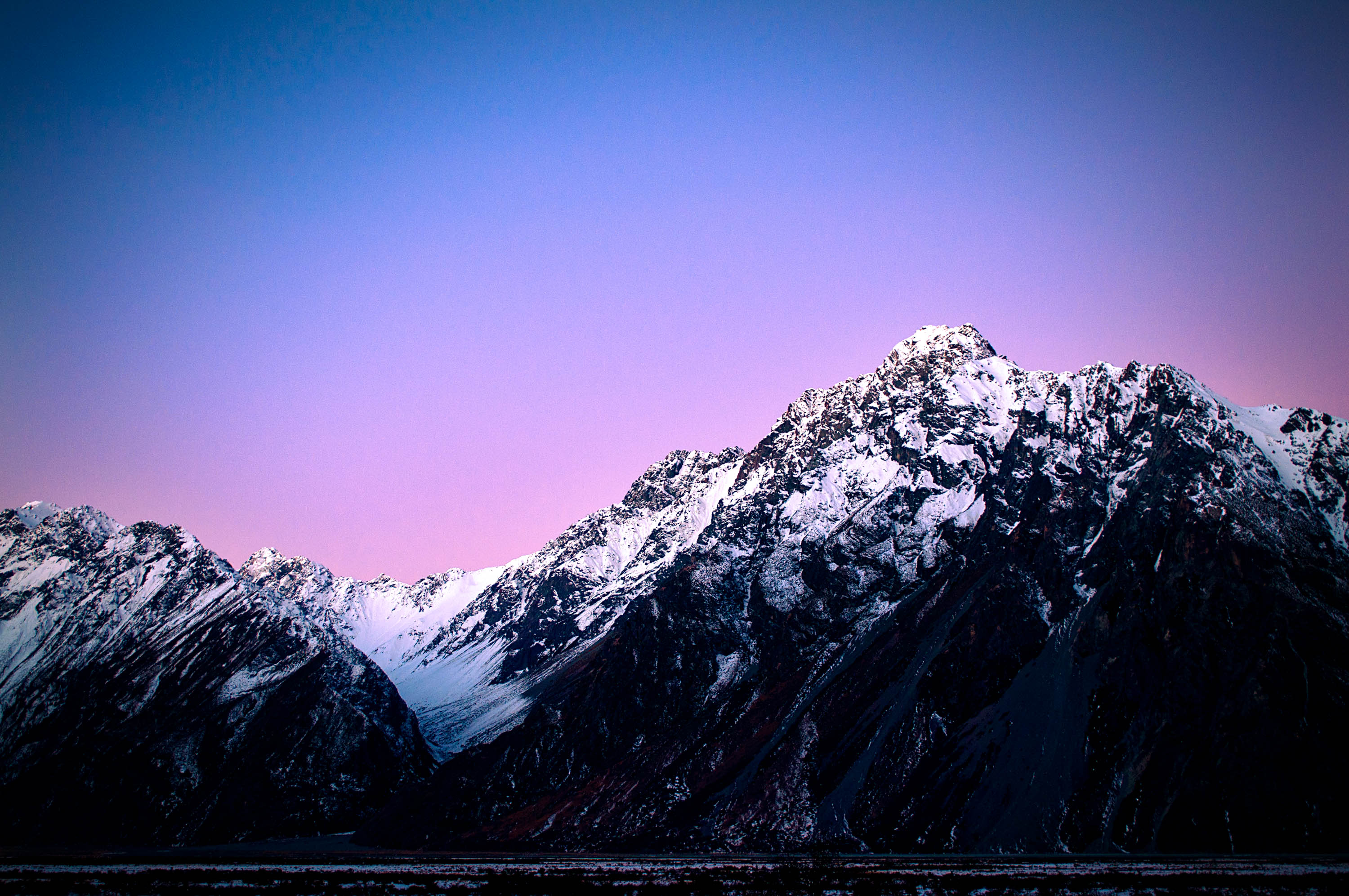 Photograph of alpine, snow-capped mountains nestled in front of the pink and purple hues of a sky at sunset, located in New Zealand.