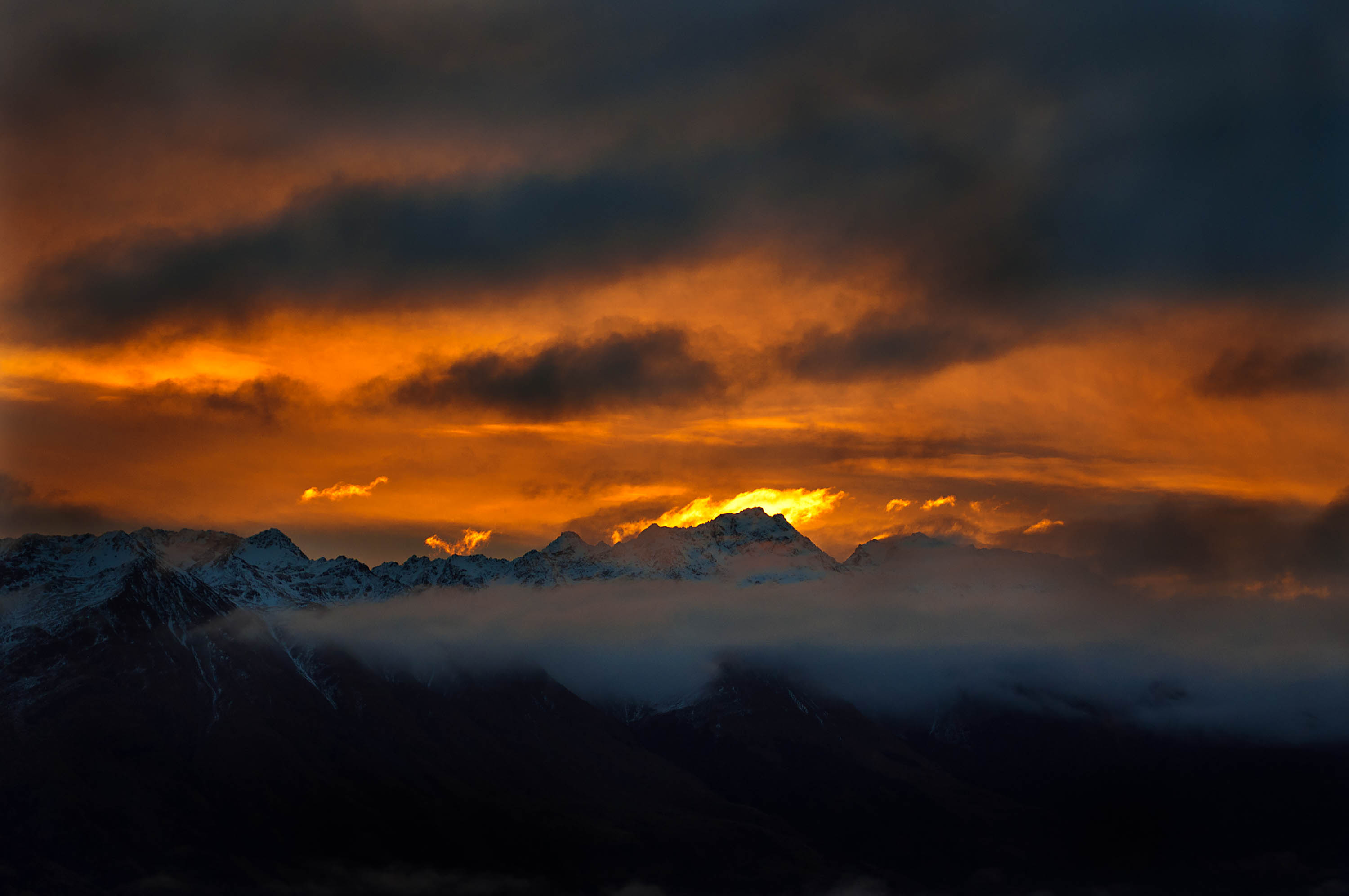 Sunset behind the mountains overlooking Lake Wakatipu, New Zealand. Long white clouds stretch out in front of the peaks, and the sky is filled with a golden-orange color, vibrantly illuminated by the light of the setting sun.