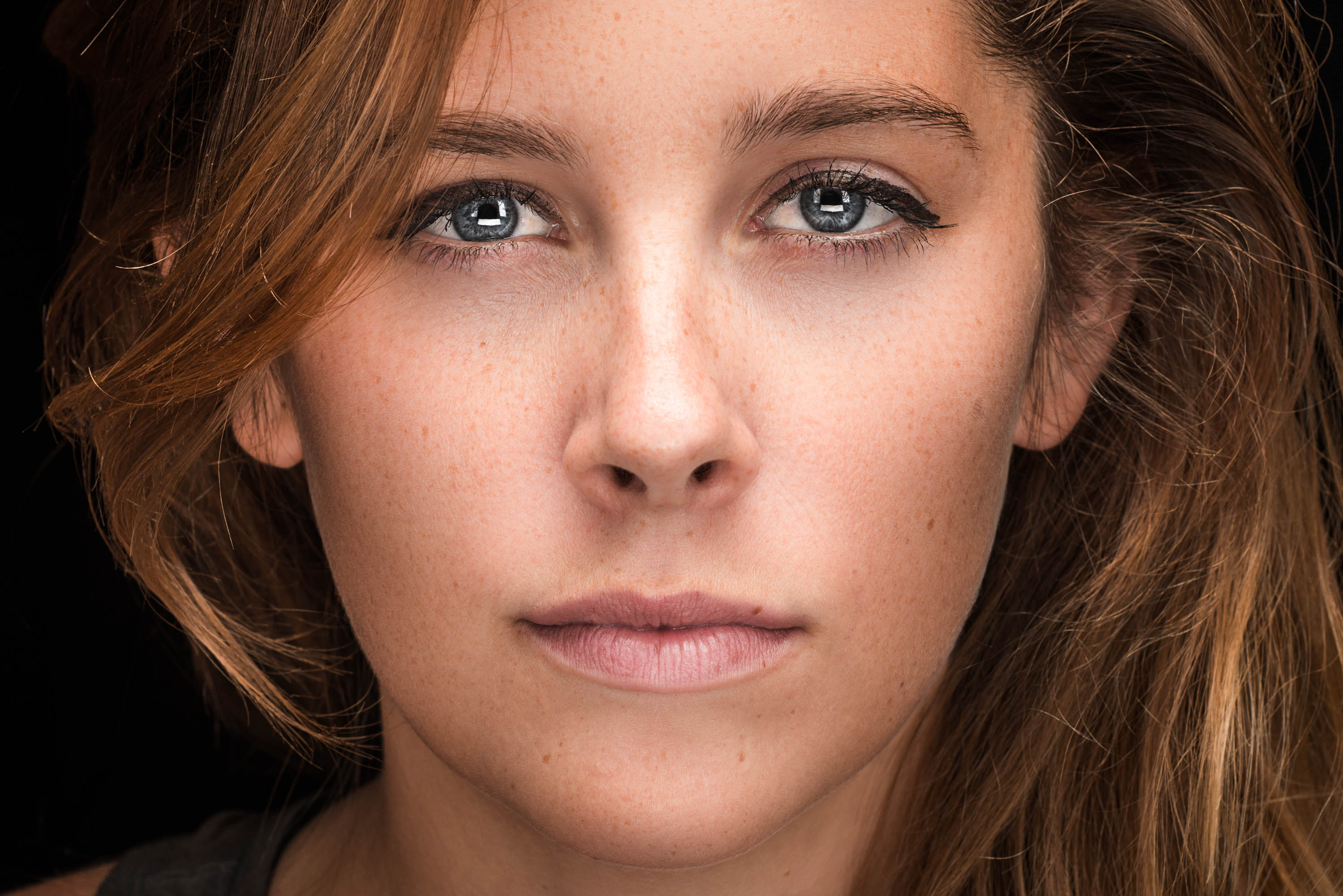 A very close-up portrait of the face of a woman's flowing brunette hair, bright catch-light in blue eyers, and freckles lining her nose and cheeks.