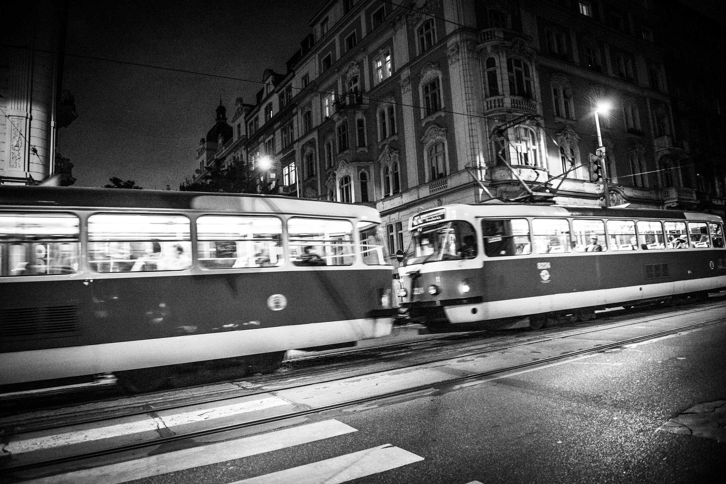 Black & white image of a city metro line with a train moving through the image under the streetlights, with buildings in the background. The movement of trains is slightly blurred.