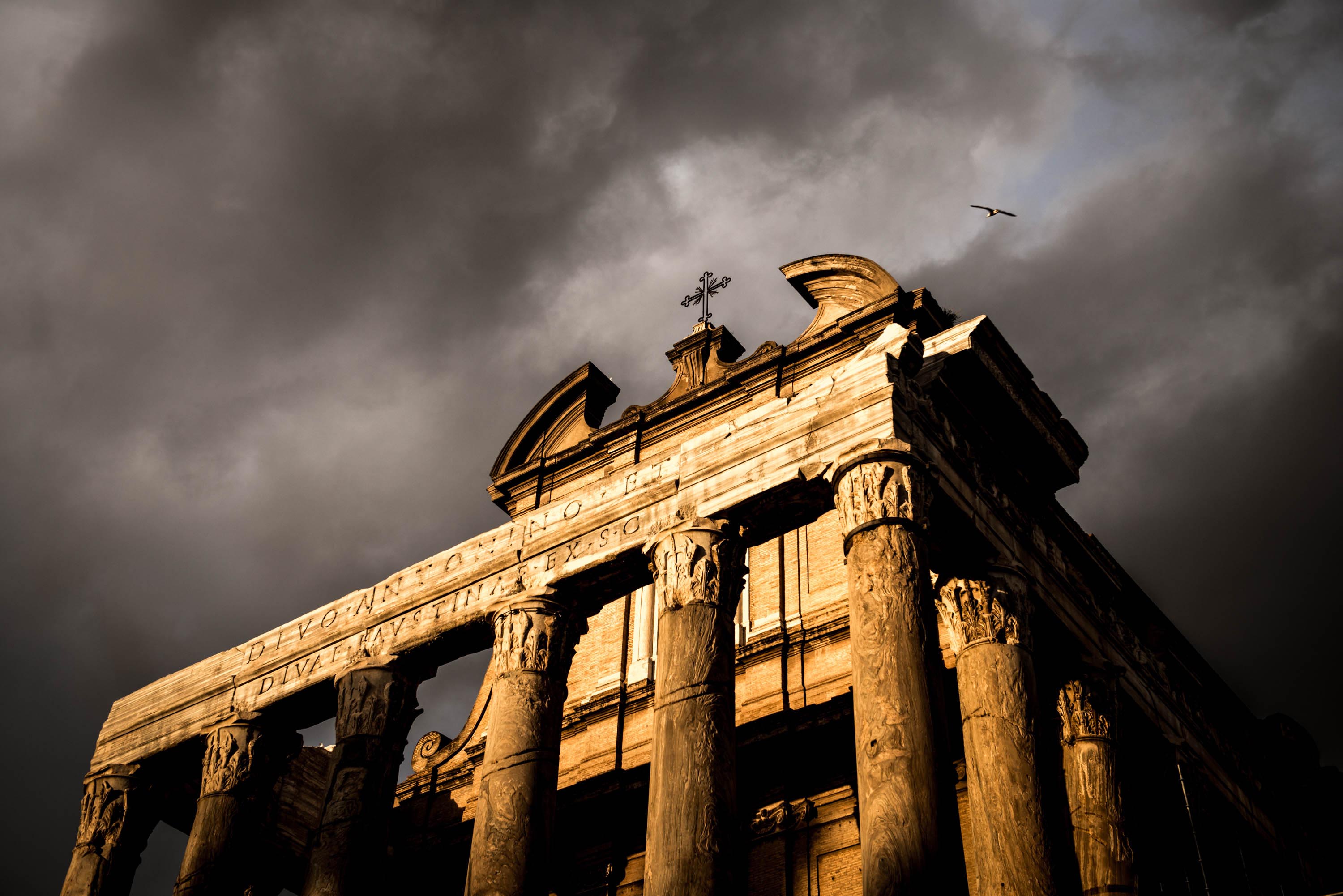 Image looking up at ancient ruins located at the Roman Forum in Italy. There is a bird flying overhead and cloud skies in the background.